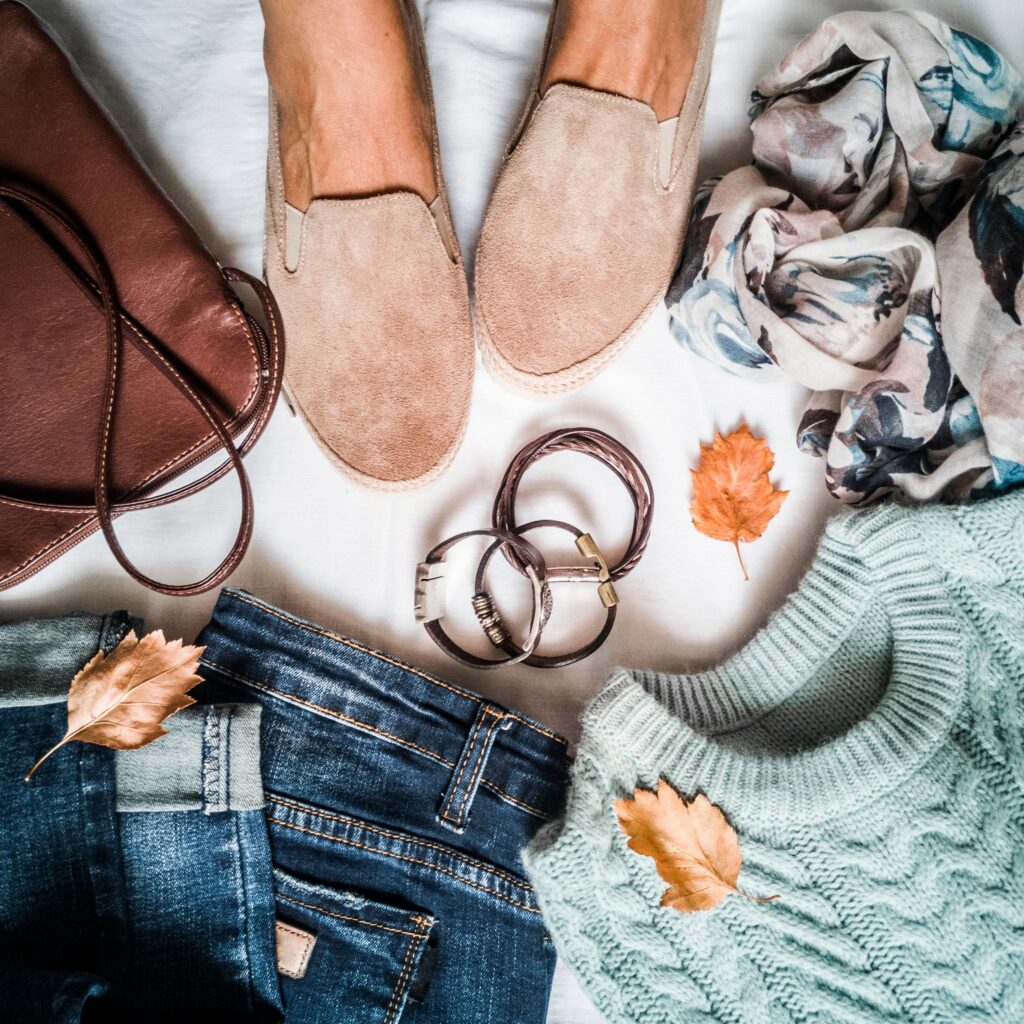 Women's autumn clothing set - jeans, suede sneakers, pullover, scarf and leather bag. Women's feet in suede sneakers surrounded by clothes for autumn walks on a light background, top view. Flat lay